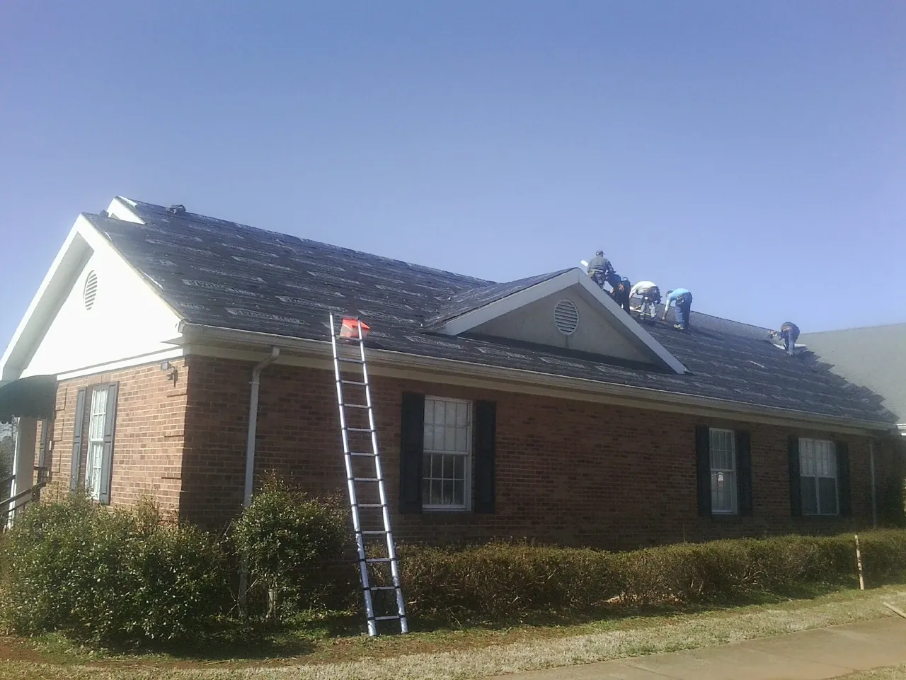 Crew tearing shingles off a brick ranch roof with a ladder set — Emergency Roof Repair work by our crew in Long Lake