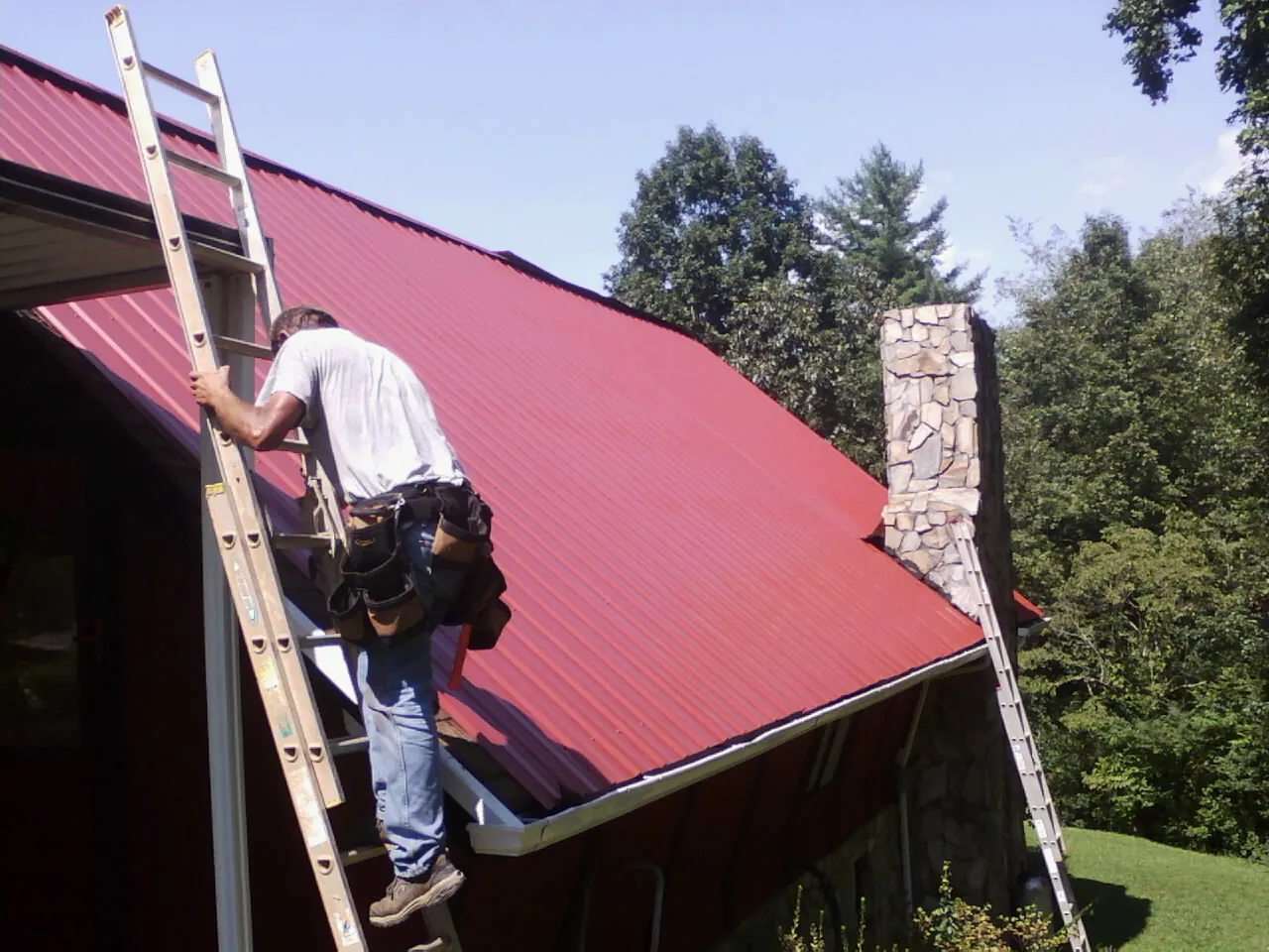 Roofer on a ladder beside a red metal roof and stone chimney — Soffit & Fascia Repair by Roofing Brothers in Long Lake
